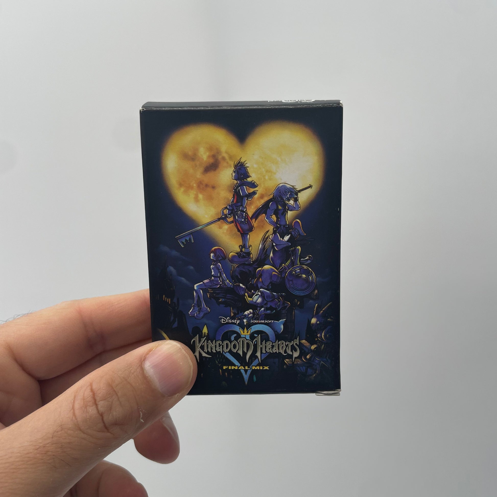 Hand holding a Kingdom Hearts trading card pack with characters and a heart design on a white background