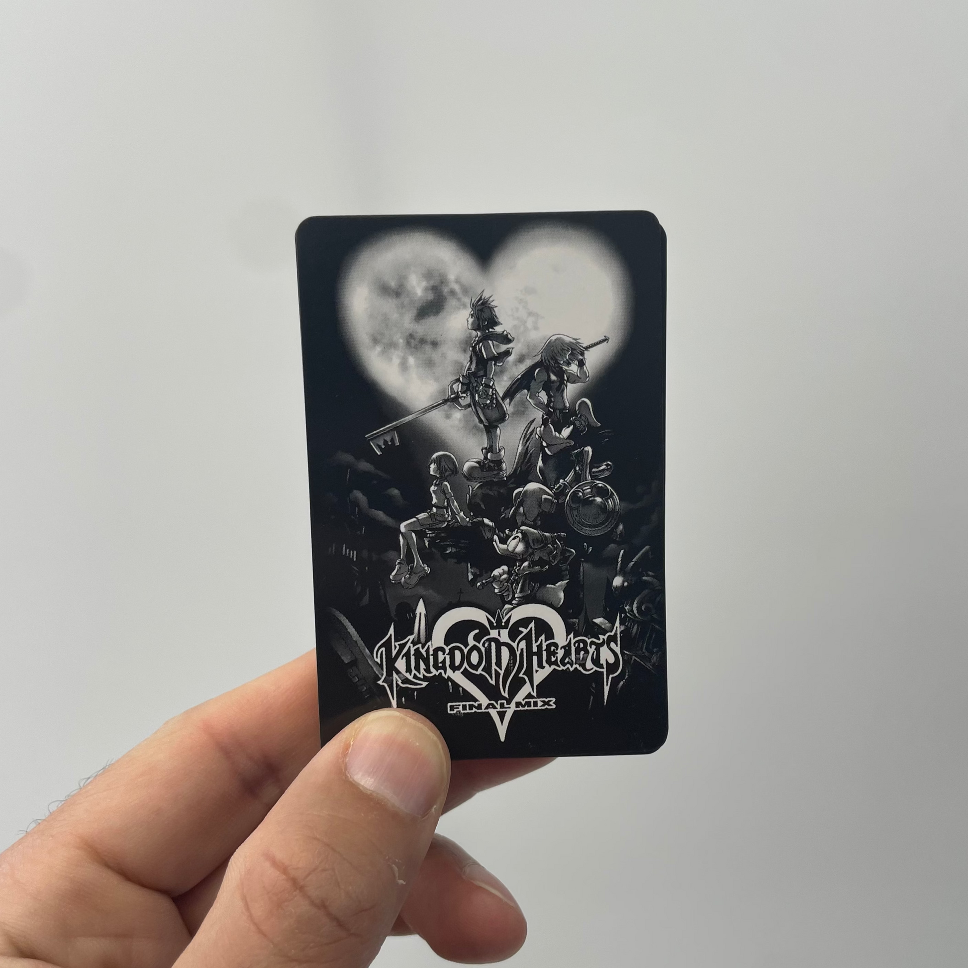 Hand holding a black card with 'Kingdom Hearts' artwork against a light gray background