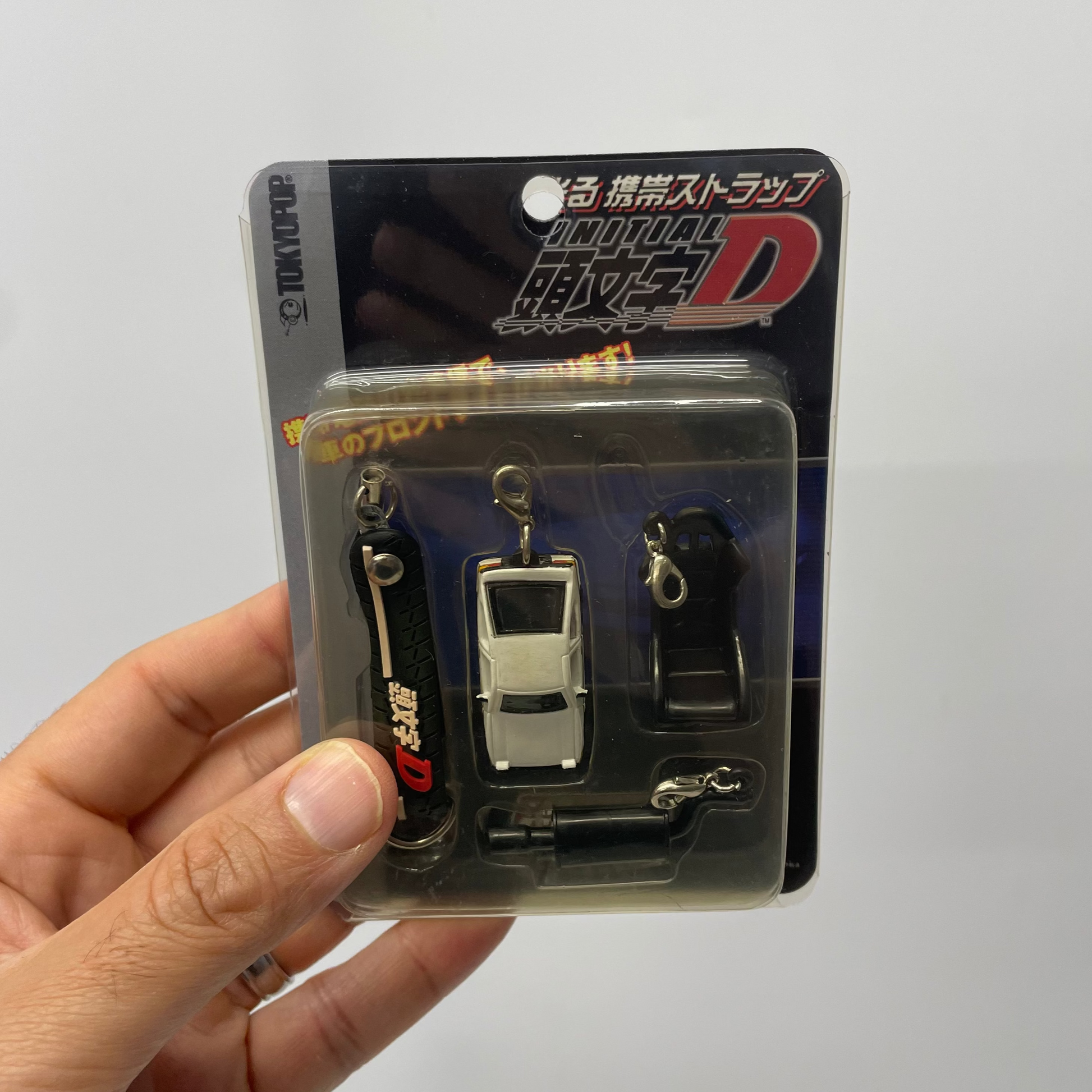 Packaged keychains with 'Initial D' branding held by a hand against a white background