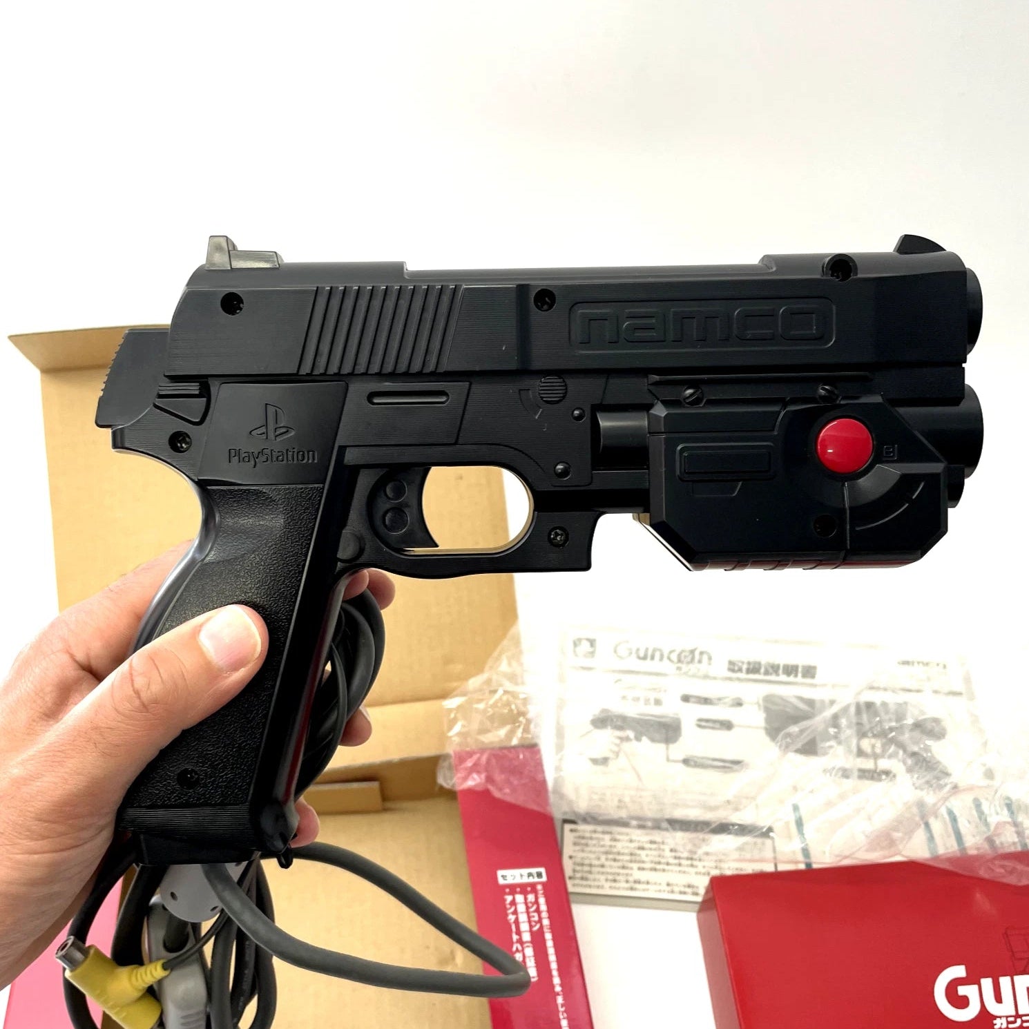Black toy gun with red button held by a hand, with packaging in the background.
