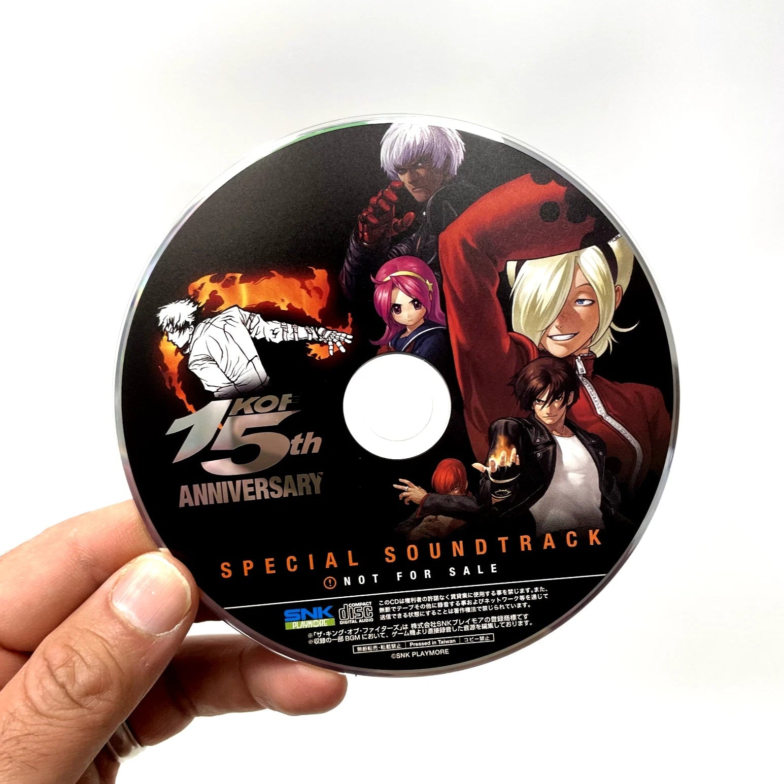 CD with anime characters and 'KOF 15th Anniversary' text held by a hand on a white background