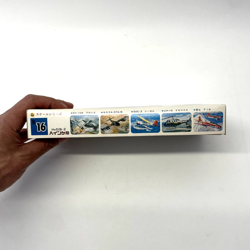 Hand holding a model airplane kit box with illustrations of various aircraft on a white background
