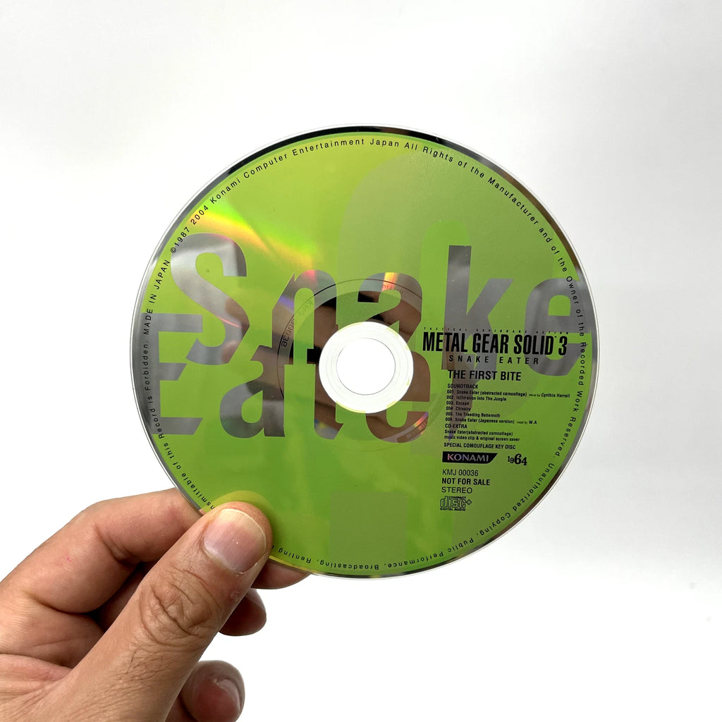 Hand holding a green 'Metal Gear Solid 3' game disc with 'Snake Eater' text.